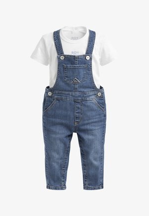 Camicia bianca a maniche corte abbinata a salopette di denim blu con tasca frontale, bretelle regolabili, bottoni laterali e chiusure a scatto lungo le gambe.