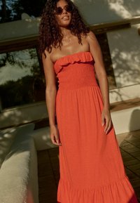 Maxime dress made of lightweight fabric in bright orange, featuring a ruffled strapless neckline and a smocked bodice, with a flared hem.