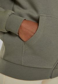 Sudadera verde olivo con puños y dobladillo acanalados, que cuenta con un bolsillo lateral y una textura de algodón suave. Detalle de cremallera no visible.