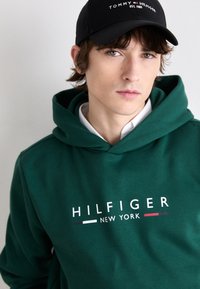 Zelena dukserica s kapuljačom i prednjim logotipom "HILFIGER NEW YORK", s mekanim teksturom. Crna kapa s bijelim natpisom i logotipom.