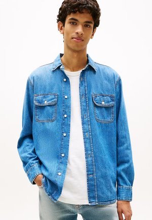 Giovane uomo con capelli ricci che indossa una camicia di jeans azzurro chiaro abbottonata sopra una t-shirt bianca, con le mani in tasca, in piedi davanti a uno sfondo semplice.