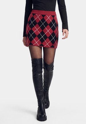 Frau trägt einen roten und schwarzen Argyle-Minirock, schwarze transparente Strumpfhosen, schwarze Overknee-Stiefel und ein schwarzes Langarmoberteil vor weißem Hintergrund.