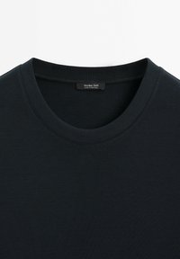 Schwarzes Rundhalsshirt mit geripptem Kragen, das den oberen Brustbereich und ein Etikett innerhalb des Halsausschnitts zeigt.