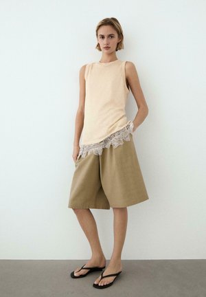 Frau steht an einer weißen Wand, trägt ein ärmelloses beiges Oberteil mit Spitzenabschluss, breite khakifarbene Shorts und schwarze Flip-Flops, Hände in den Taschen.