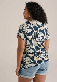 Top a maniche corte in un motivo a foglie blu, beige e crema. Tessuto morbido e leggero con maniche arrotolate. Abbinato a pantaloni corti in denim.