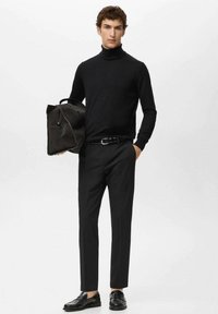 Zwarte coltrui, donkere broek en loafers. Draagt een donkere, gestructureerde tas met een handvat bovenop, met een minimalistisch ontwerp en accenten.