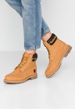 Personne portant un jean bleu clair et des bottes Timberland marron clair à lacets, debout sur un sol gris clair avec un pied légèrement levé.