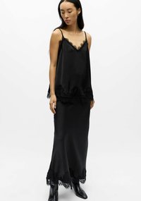 Camisole en satin noir avec bordure en dentelle, associée à une longue jupe en satin noir ornée d'accents de dentelle à l'ourlet, portée avec des bottines noires.