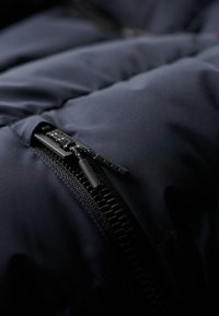 Puffjakke i mørk marineblå, med et nærbillede af en sort lynlås med teksturerede trækknapper og en glat, mat stofoverflade.