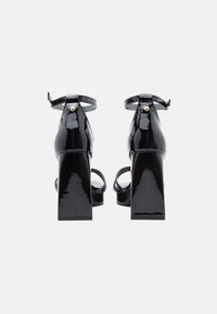 Fekete lakkbőr magas sarkú szandál vastag sarokkal, bokapánttal és fényes felülettel, a pánton egy kis arany díszítéssel.