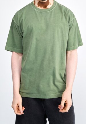 Person mit Vitiligo, die ein schlichtes grünes T-Shirt mit kurzen Ärmeln und dunkle Hosen trägt, vor einem weißen Hintergrund stehend.