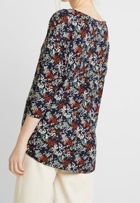 Kwiatowa bluzka z rękawami 3/4, granatowe tło z czerwonymi, zielonymi i białymi wzorami liści, miękki materiał, zaokrąglony dół, zestawiona z jasnymi spodniami.