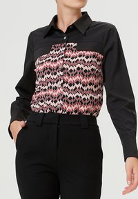 Chemise noire à manches longues avec un devant à motif géométrique en rose, rouge et noir, fermeture à boutons et col structuré.