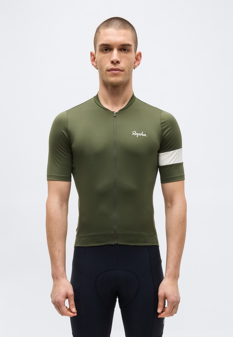 Άνδρας μοντέλο φοράει ελαιί πράσινη κοντομάνικη ποδηλατική φανέλα με λευκή λωρίδα στο μανίκι και μαύρο επενδυμένο ποδηλατικό σορτς, στέκεται μπροστά σε απλό φόντο.