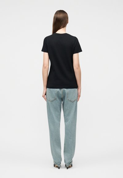 Γυναίκα που στέκεται με την πλάτη γυρισμένη, φοράει μαύρο κοντομάνικο μπλουζάκι, ανοιχτόχρωμα φαρδιά τζιν και μαύρες μυτερές γόβες, σε λευκό φόντο.