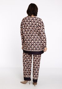Vrouw gezien van achteren in een bijpassend lang mouwtop en broek met een geometrisch druppelpatroon in zwart, roze en beige, met beige hakken.