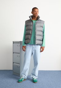 Grå puffervest, grønstribet langærmet skjorte, lyseblå jeans, grønne sneakers med hvide striber, stående ved siden af et gråt arkivskab.