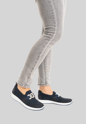 Beine in hellgrauen Skinny-Jeans und marineblauen Slipper-Sneakern mit weißen Sohlen und silbernem Schnallendetail vor einem einfarbigen grauen Hintergrund.