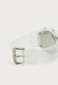 Gennemsigtig silikone urearmbånd med en sølvmetal spænde, der har en glat tekstur og et minimalistisk design. Bagsiden har en metalfinish.