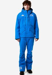 Giacca impermeabile blu con cappuccio, due tasche anteriori con zip e dettagli con logo. Abbinata a pantaloni blu coordinati e calzature nere.