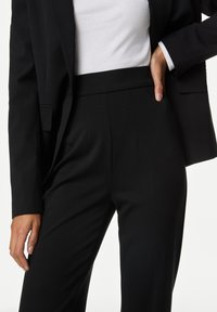 Schwarze maßgeschneiderte Hose mit glatter Textur, mit Falten und hoher Taille. Kombiniert mit einem weißen Oberteil und einem schwarzen Blazer.