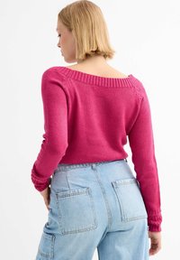 Suéter de punto rosa ajustado con un escote hombro caído, detalles acanalados en los puños y el cuello, combinado con unos jeans de cintura alta en azul claro.