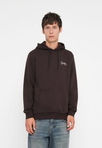 Mørkebrun hættebluse med snore, frontlomme og hvid broderet logo. Båret med lyseblå jeans, der har en afslappet pasform.