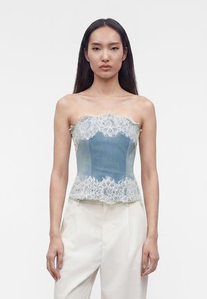 Kobieta o długich ciemnych włosach, ubrana w niebieski jeansowy gorset bez ramiączek z białą koronkową lamówką oraz białe, wysokie spodnie, stojąca na jednolitym tle.