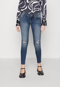 Jeans ajustados en denim azul oscuro con áreas desgastadas, con cinturilla de talle medio y detalle de costuras mínimas. Combinados con zapatos negros.