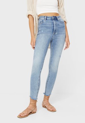 Frau trägt hellblaue Skinny-Jeans mit hoher Taille und ausgefransten Säumen, kombiniert mit beigefarbenen Schnürsandalen und einem weißen Cropped-Top sowie einem beigen Hemd.