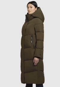 Vrouw in een lange, donkerolijfgroene gewatteerde winterjas met hoge kraag, capuchon en ritszakken aan de zijkant, staande tegen een effen achtergrond.