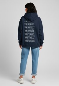 Felpa blu navy con testo bianco che elenca frasi relative all'arte e alla cultura sul retro. Abbinate a pantaloni azzurri chiari e scarpe bianche.