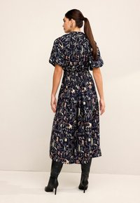 Rochie bleu marin de lungime medie, până la glezne, cu talie încrețită, mâneci bufante și un model geometric cu culori deschise; purtată cu cizme negre.