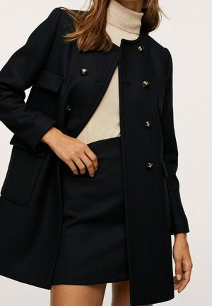 Femme portant un manteau noir croisé sur un col roulé beige et une jupe noire, une main posée sur la taille.