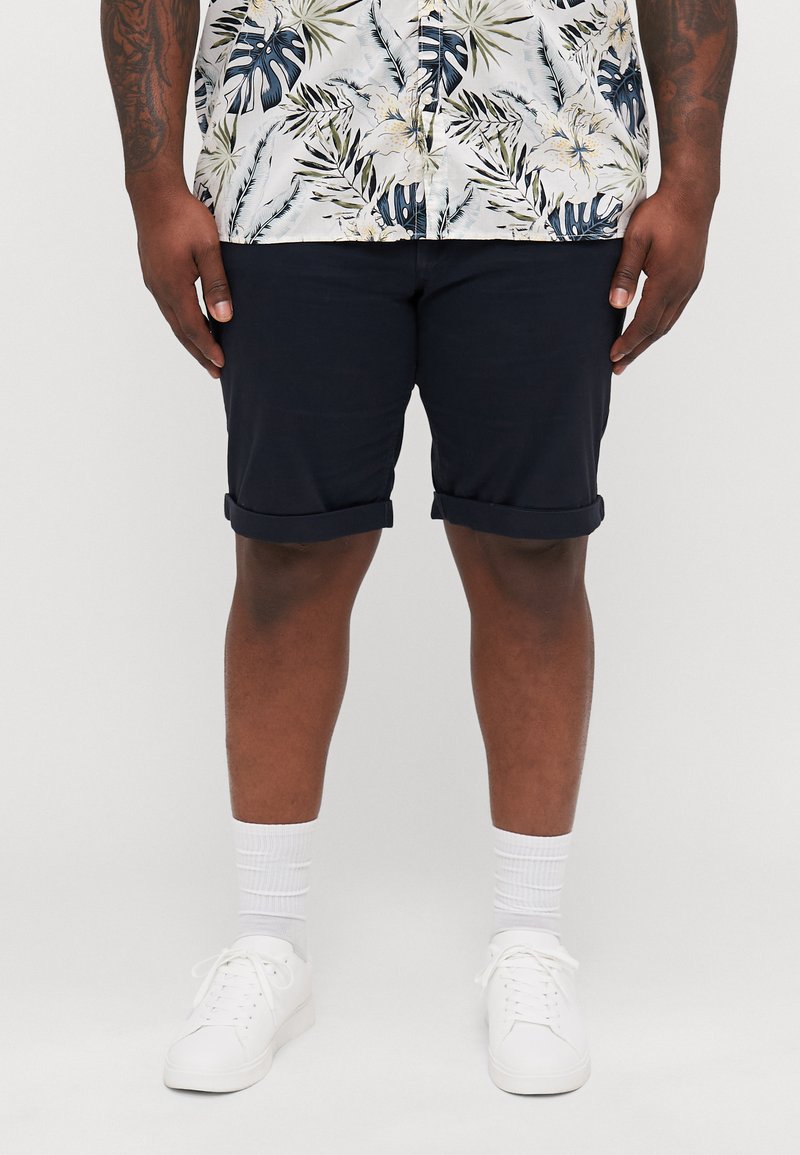 Pantalones cortos de marinero con dobladillos enrollados, combinados con una camisa de patrón tropical que presenta diseños florales en verde, azul y blanco. Zapatillas blancas y calcetines.