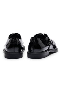 Chaussures en cuir verni noir avec une pointe arrondie, un talon légèrement surélevé et une semelle en caoutchouc noir texturé. Présente un logo sur le talon.