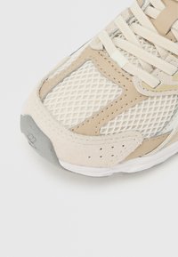 Beige és törtfehér sportcipő orr részének közeli képe, amely hálós anyagot, velúr- és bőrrészleteket, valamint fehér fűzőket tartalmaz, tiszta fehér háttéren.