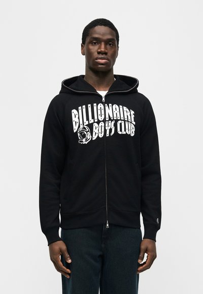 Νεαρός άνδρας που φοράει μαύρο φούτερ με φερμουάρ, με το λογότυπο "Billionaire Boys Club" και γραφικό με κράνος αστροναύτη στο μπροστινό μέρος, μπροστά σε απλό φόντο.