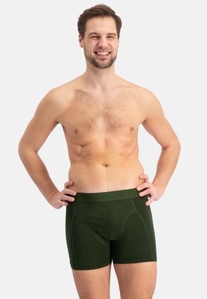 Lachende man staand met handen in de zij, gekleed in donkergroene boxershorts, tegen een eenvoudige lichte achtergrond.