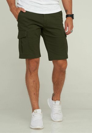 Shorts cargo vert olive en coton, avec poches latérales, longueur aux genoux et coupe droite ; portés avec des baskets blanches.