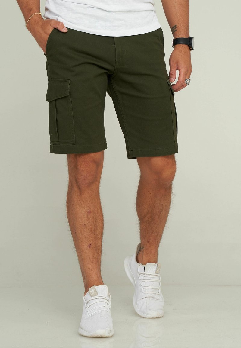 Shorts cargo vert olive en coton, avec poches latérales, longueur aux genoux et coupe droite ; portés avec des baskets blanches.