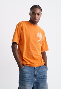 Orange bomulds t-shirt med rund hals og korte ærmer. Har et hvidt grafisk design på forsiden. Bæres med blå denim jeans.