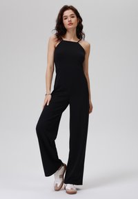 Schwarzer Jumpsuit mit Spaghettiträgern, weitem Bein und tailliertem Oberteil. Das Model trägt weiße Sneakers mit braunen Akzenten. Schlichter Hintergrund.