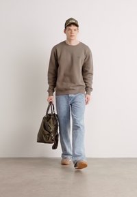 Oliven grønn hettegenser, lyseblå jeans, lysebrune sko, bærer en mørkegrønn veske. Enkelt design med en enkel logo på genseren.