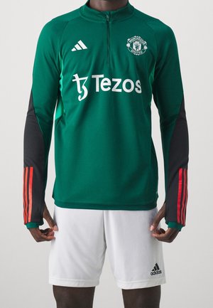 Personne porte un haut d’entraînement vert de Manchester United avec le sponsor Tezos, un logo adidas, des manches noires à bandes rouges et un short adidas blanc.