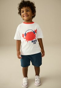 T-shirt en coton blanc avec un col rouge accentué, présentant un graphique d'un crabe rouge portant des écouteurs et le texte "AMUSEZ-VOUS", associé à un short marine et des chaussures blanches.