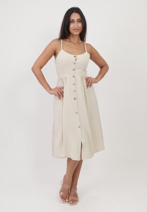 Abito midi beige con spalline sottili, chiusura frontale con bottoni, vita arricciata, tessuto testurizzato e uno spacco all'orlo, abbinato a sandali con tacco trasparente.