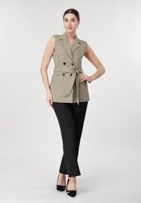 Γυναίκα στέκεται φορώντας αμάνικο μπεζ γιλέκο με ζώνη, μαύρο παντελόνι και μαύρα τακούνια μπροστά σε απλό φωτεινό φόντο.