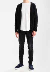 Man står klädd i en vit t-shirt, svart öppen kofta, svarta jeans och marinblå sneakers framför en helt vit bakgrund.