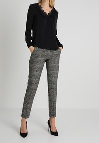 Schwarze Bluse mit langen Ärmeln und Spitzendetail am Ausschnitt, kombiniert mit grauen karierter Hose und schwarzen spitzen Pumps.